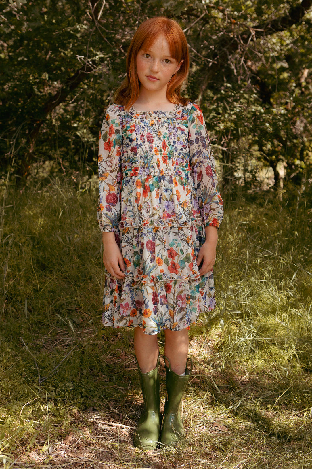 A young girl with red hair stands on grass among trees, wearing the Ivy City Co Mini Selena Dress in Vivid Meadow with a smocked bodice and green rain boots.