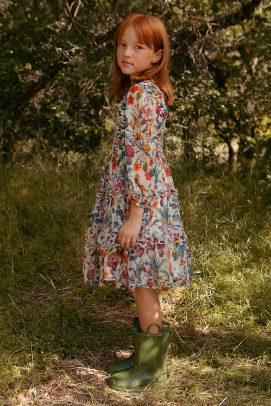 A young red-haired girl stands sideways on grass outdoors, wearing the Ivy City Co Mini Selena Dress in Vivid Meadow and green rubber boots. Lush trees and foliage form the backdrop.