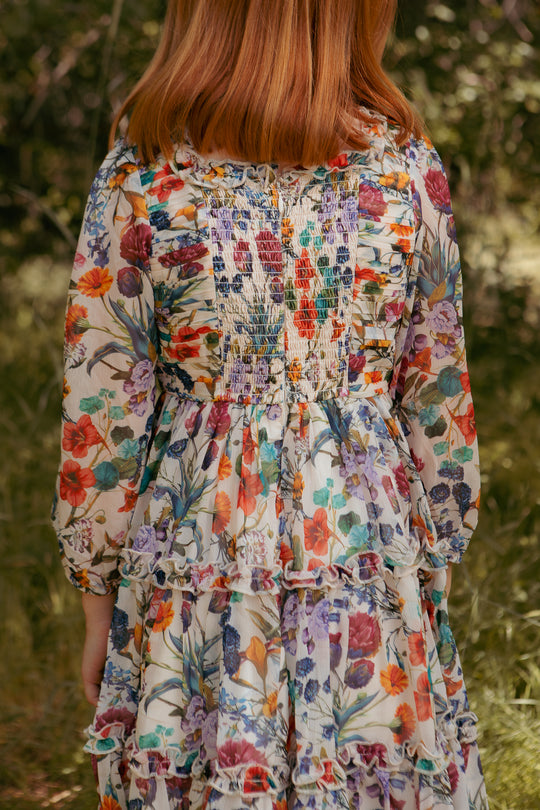 A person with reddish hair stands outdoors, shown from behind, wearing the Ivy City Co Mini Selena Dress in Vivid Meadow—a girls floral midi dress—surrounded by greenery.