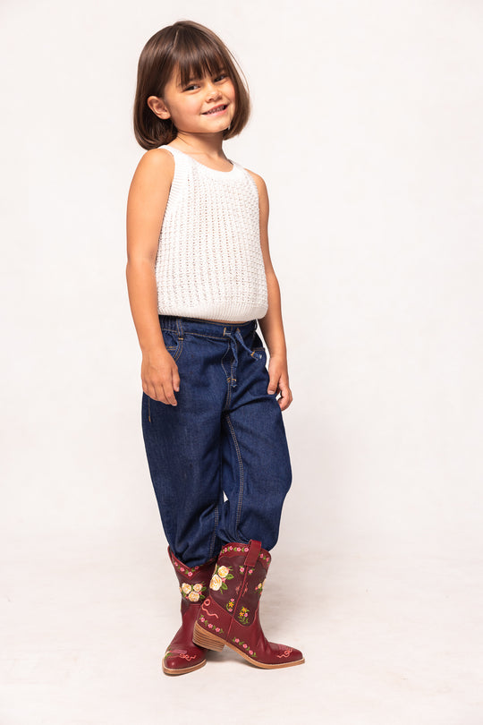 Child wearing a white sleeveless top, blue jeans, and red boots with gold patterns on a white background