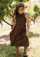 A young girl with long brown hair stands outdoors in sunlight, wearing the Ivy City Co Teen Simone Dress in Warm Cocoa—a sleeveless dress featuring a smocked bodice and handy pockets—paired with dark shoes, with trees and grass in the background.
