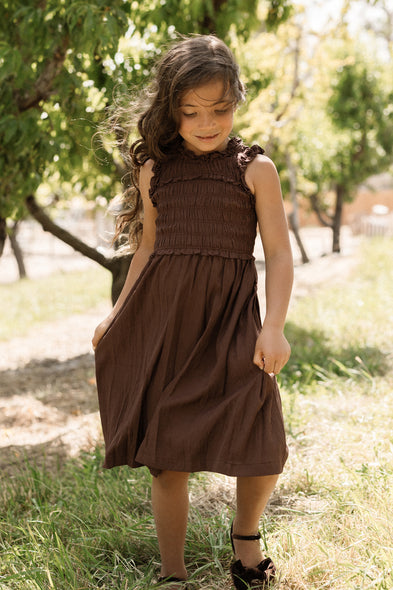 A young girl with long brown hair stands outdoors in sunlight, wearing the Ivy City Co Teen Simone Dress in Warm Cocoa—a sleeveless dress featuring a smocked bodice and handy pockets—paired with dark shoes, with trees and grass in the background.