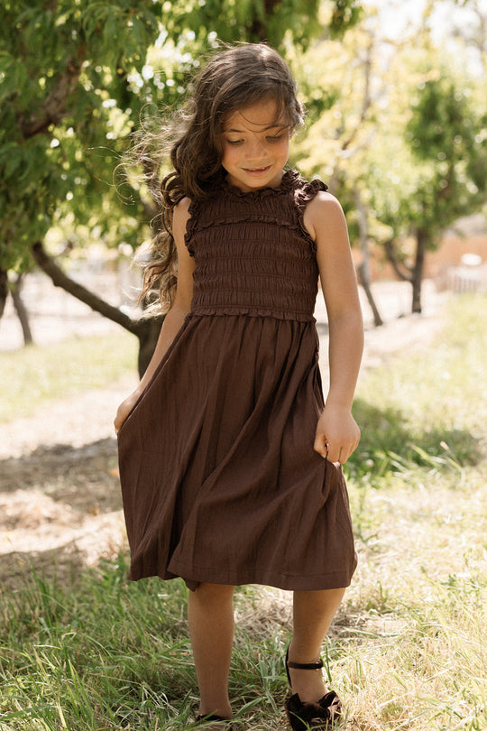 A young girl with long brown hair stands outdoors in sunlight, wearing the Ivy City Co Teen Simone Dress in Warm Cocoa—a sleeveless dress featuring a smocked bodice and handy pockets—paired with dark shoes, with trees and grass in the background.