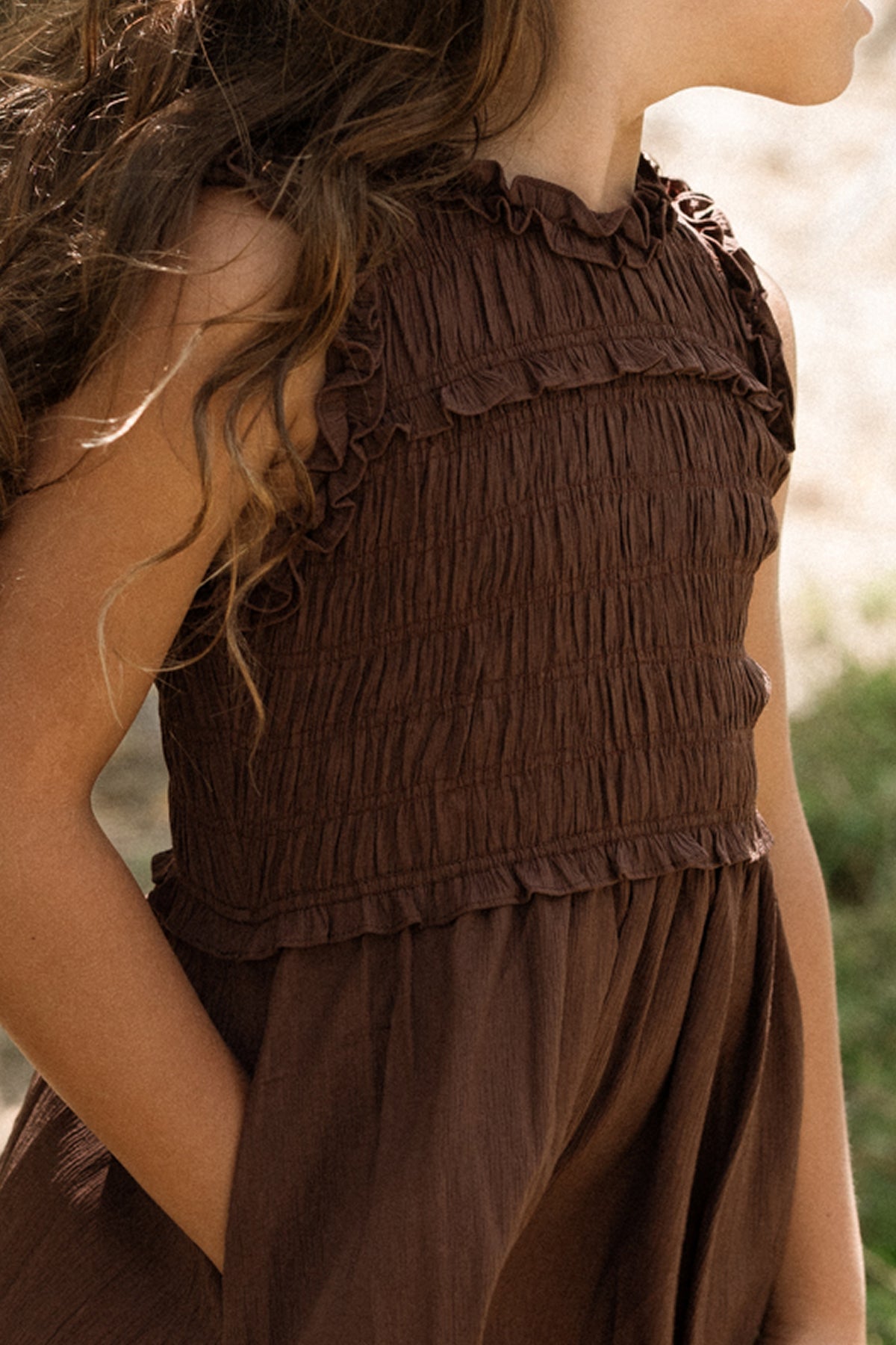 A young girl stands outdoors with one hand in her pocket, wearing the Ivy City Co Teen Simone Dress in Warm Cocoa. The sleeveless dress features a smocked bodice, and her long, wavy hair flows down her back.