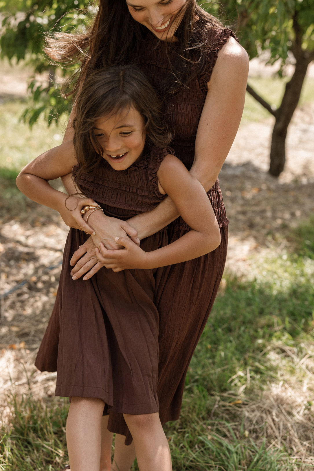 A woman hugs a smiling child from behind as they stand outside in a grassy area, both wearing matching Ivy City Co Teen Simone Dresses in Warm Cocoa, featuring smocked bodices and handy pockets.
