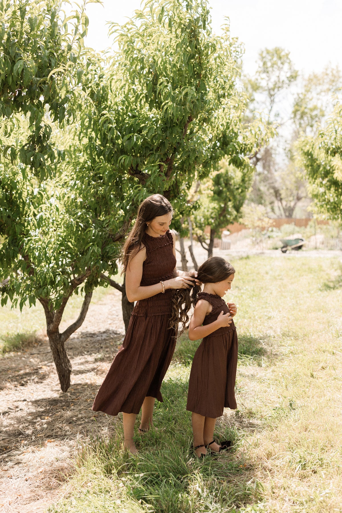 A woman braids a young girl’s hair as they stand together outdoors among green trees on a sunny day. The young girl wears the Ivy City Co Teen Simone Dress in Warm Cocoa, featuring a smocked bodice and convenient pockets, perfectly paired with sandals.