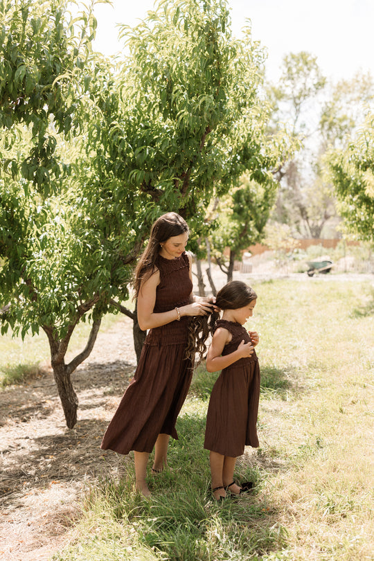 A woman braids a young girl’s hair as they stand together outdoors among green trees on a sunny day. The young girl wears the Ivy City Co Teen Simone Dress in Warm Cocoa, featuring a smocked bodice and convenient pockets, perfectly paired with sandals.