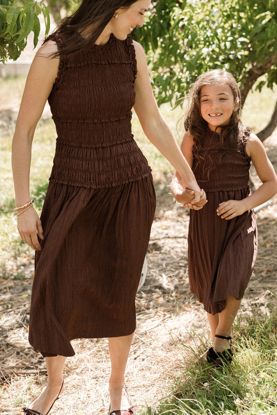 A woman and a young girl—both dressed in Ivy City Co’s Teen Simone Dress in Warm Cocoa, featuring smocked bodices and handy pockets—hold hands and smile while walking outdoors among green trees.