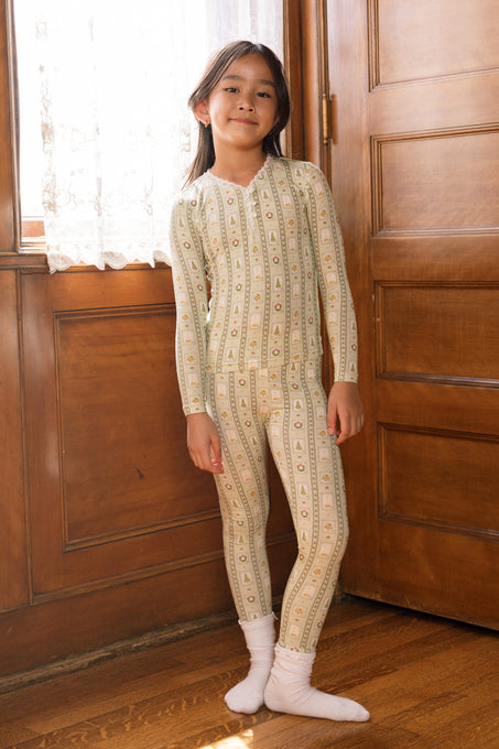 A young child smiles by wooden doors and a window, wearing the Ivy City Co Mini So Soft Henley Pajama Set in Merry Mail Linear and white socks as sunlight shines through lace curtains.