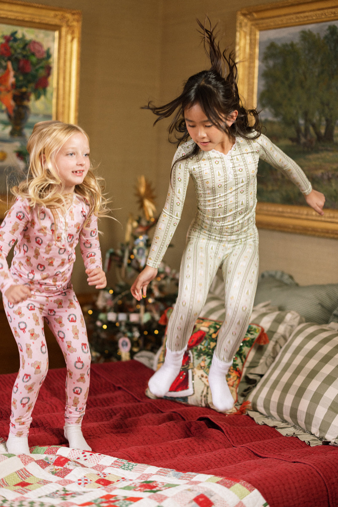 Two young children in the Mini So Soft Henley Pajama Set in Merry Mail Linear by Ivy City Co happily jump on a bed, with a Christmas tree and holiday décor creating a festive scene.
