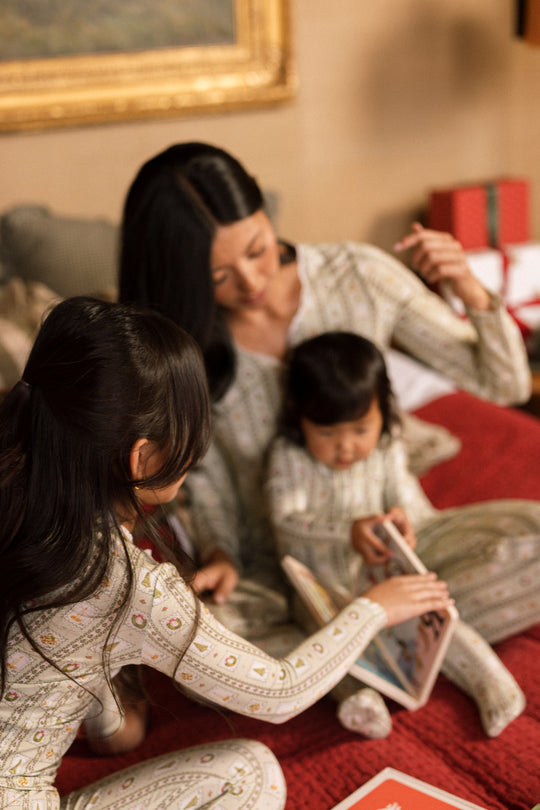 A woman and two young girls wear the Mini So Soft Henley Pajama Set in Merry Mail Linear by Ivy City Co as they read together on a bed, surrounded by wrapped gifts for a warm, festive scene.