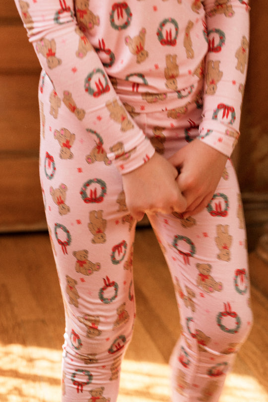 A child in the Mini So Soft Henley Pajama Set in Teddy Bear Wreaths by Ivy City Co stands indoors on a wooden floor, hands clasped, ready for cozy family matching moments.
