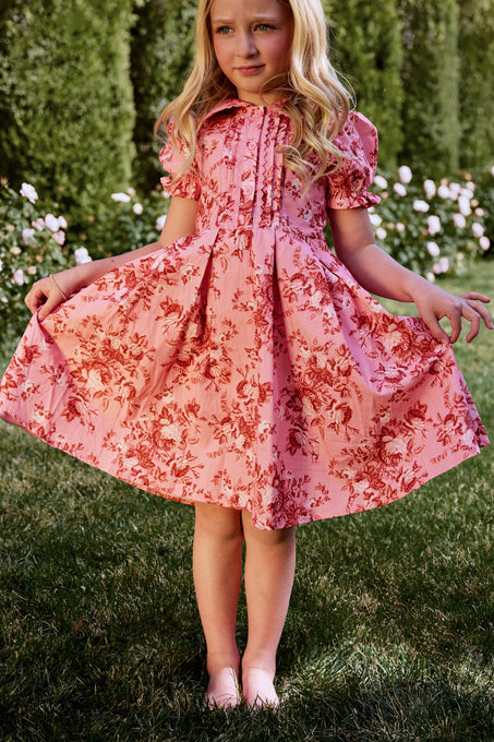 A young girl wearing the Mini Sophia Dress in Desert Heritage Rose by Ivy City Co stands barefoot outdoors, holding out her skirt. She has long blonde hair, with green bushes and white flowers in the background.