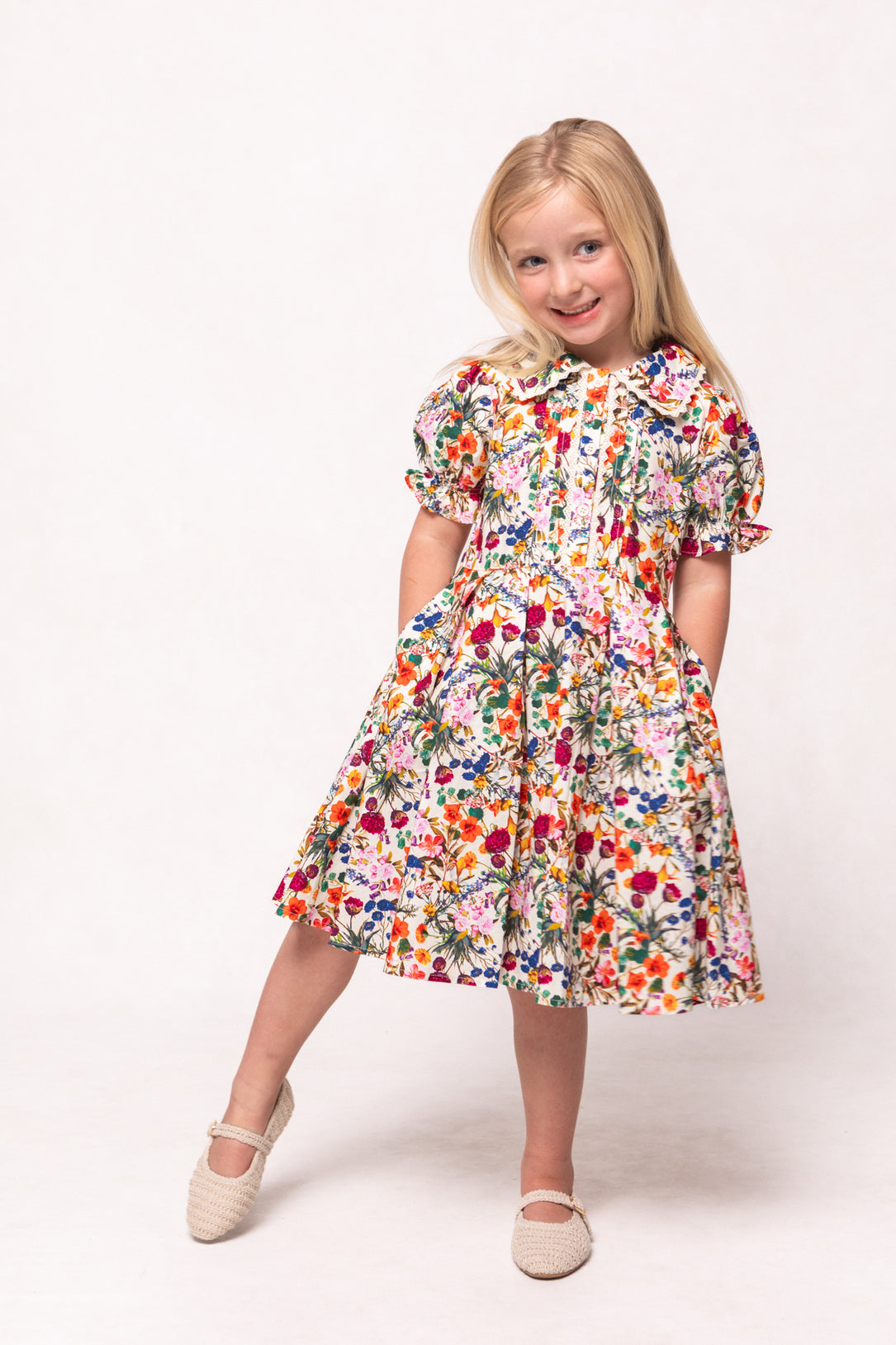 A young girl with long blonde hair smiles, wearing the Mini Sophia Dress in Multicolor Floral with puffed sleeves and white shoes, standing against a plain white background.