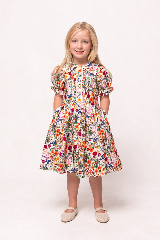 Young girl with long blonde hair wears the Ivy City Co Mini Sophia Dress in Multicolor Floral, standing with hands in her pockets and smiling against a plain white background.