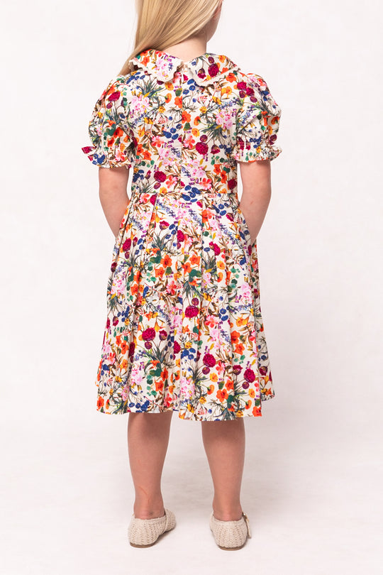 A young girl with light hair faces away, wearing the Ivy City Co Mini Sophia Dress in Multicolor Floral—a knee-length, short-sleeved dress with a full skirt and puffed sleeves—paired with light woven shoes.