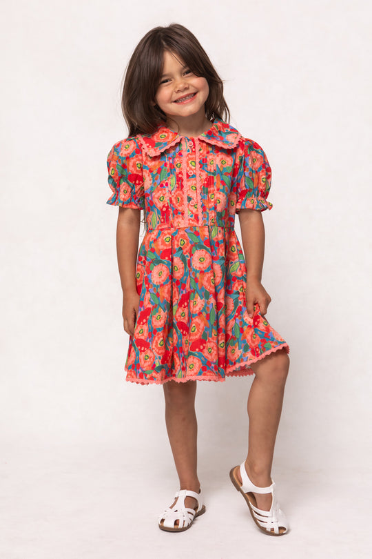 A young girl with long brown hair smiles while wearing the Mini Sophia Dress in Poppy, featuring puffed sleeves and a pleated skirt. She stands on one leg against a plain white background, pairing the dress with white sandals.