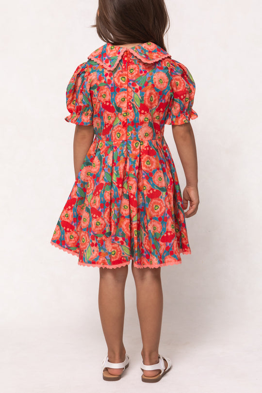 A young girl with brown hair stands facing away, wearing the Mini Sophia Dress in Poppy—a colorful cotton dress with puffed sleeves, a scalloped collar, pleated skirt—and white sandals.