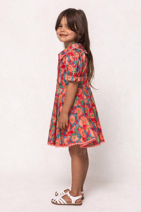 A young girl with long brown hair smiles in the Ivy City Co Mini Sophia Dress in Poppy, featuring puffed sleeves and a pleated skirt, paired with white sandals against a plain light background.