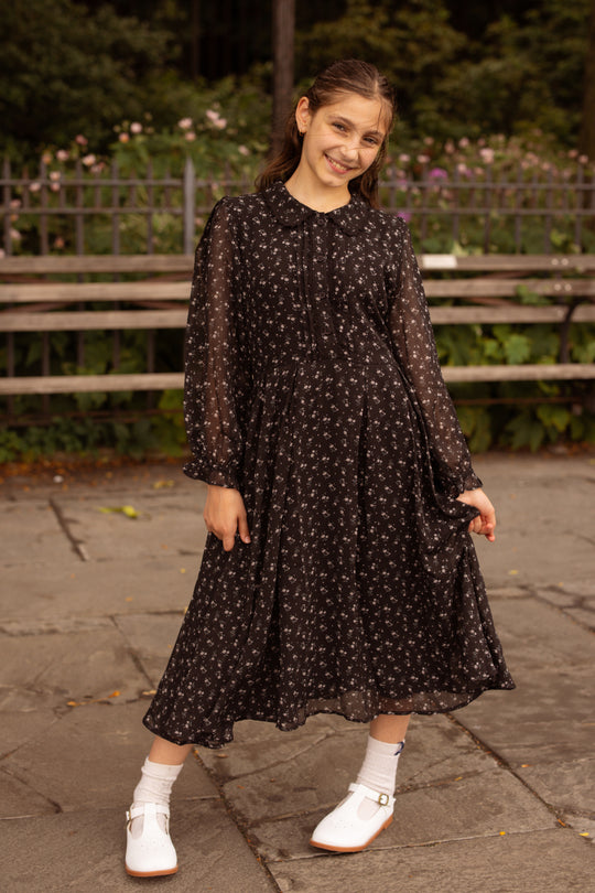 A young girl smiles while standing outdoors on stone pavement. She wears the Ivy City Co Mini Sophia Long Sleeve Dress in Noir Ditsy Chiffon, paired with white shoes and socks. A wooden bench and greenery are in the background.