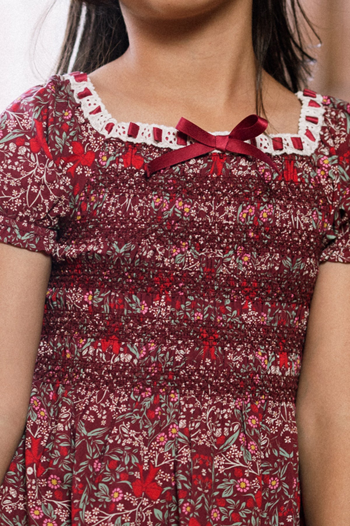 A young girl wearing the Ivy City Co Mini Sunday Best Dress in Garnet Ribboned Garden, featuring a lace-trimmed neckline and a small red bow at the collar. The image is cropped to show her from shoulders to waist.