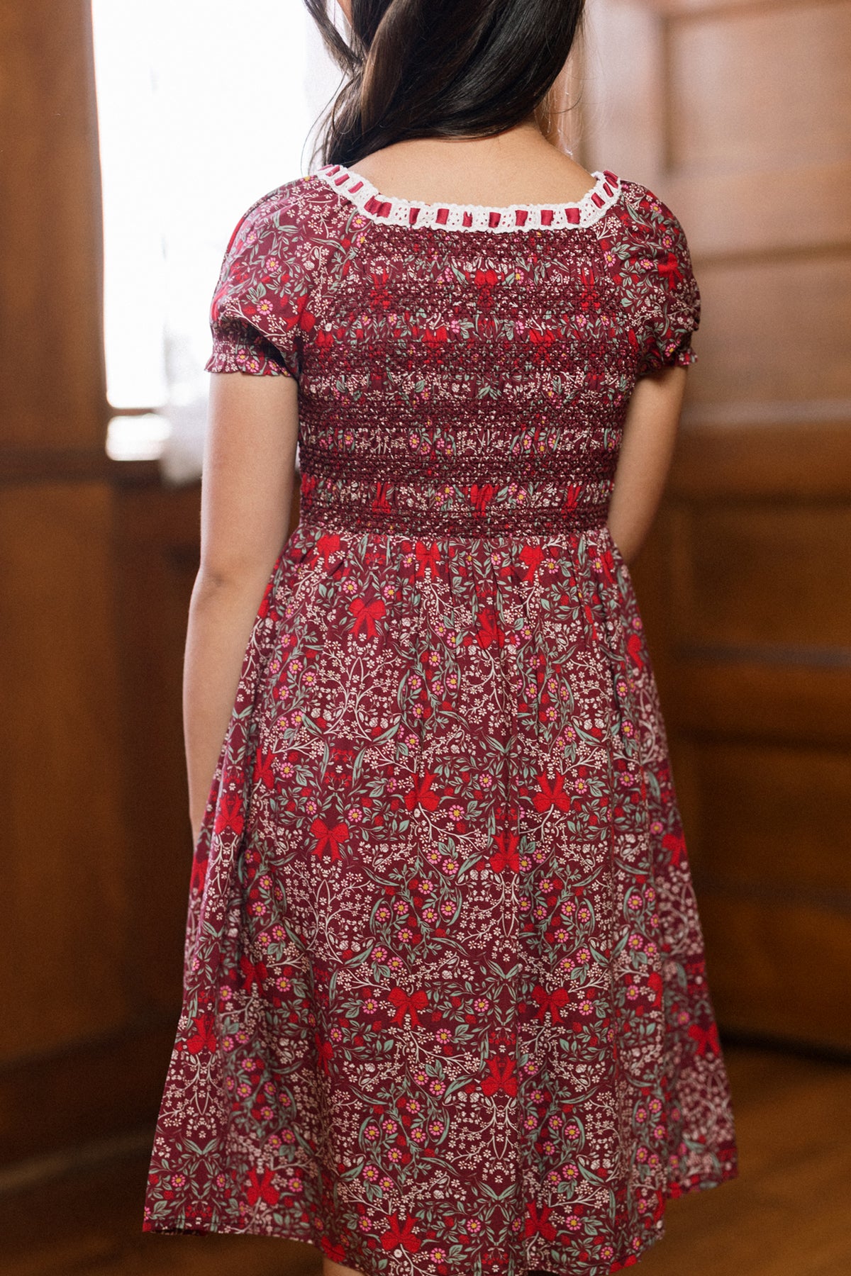 A woman with long dark hair, seen from behind, wears the Ivy City Co Mini Sunday Best Dress in Garnet Ribboned Garden, featuring a smocked bodice and short sleeves, as she stands indoors by a wooden wall.