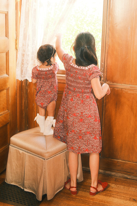 Two young girls in matching Ivy City Co Mini Sunday Best Dresses in Garnet Ribboned Garden gaze out a sunlit window. The younger child, wearing this charming holiday dress featuring a delicate smocked bodice, stands atop an ottoman, while the older girl stands beside her—both facing away in the warmly lit wooden room.