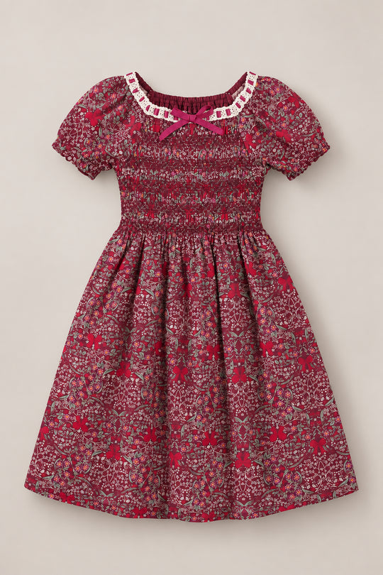 The Mini Sunday Best Dress in Garnet Ribboned Garden by Ivy City Co is a short-sleeved girls’ holiday dress showcasing a vibrant garnet floral pattern, with a smocked bodice, puff sleeves, white lace neckline trim, and a delicate red bow at the center. Its gathered skirt flows outward for an effortlessly charming look.