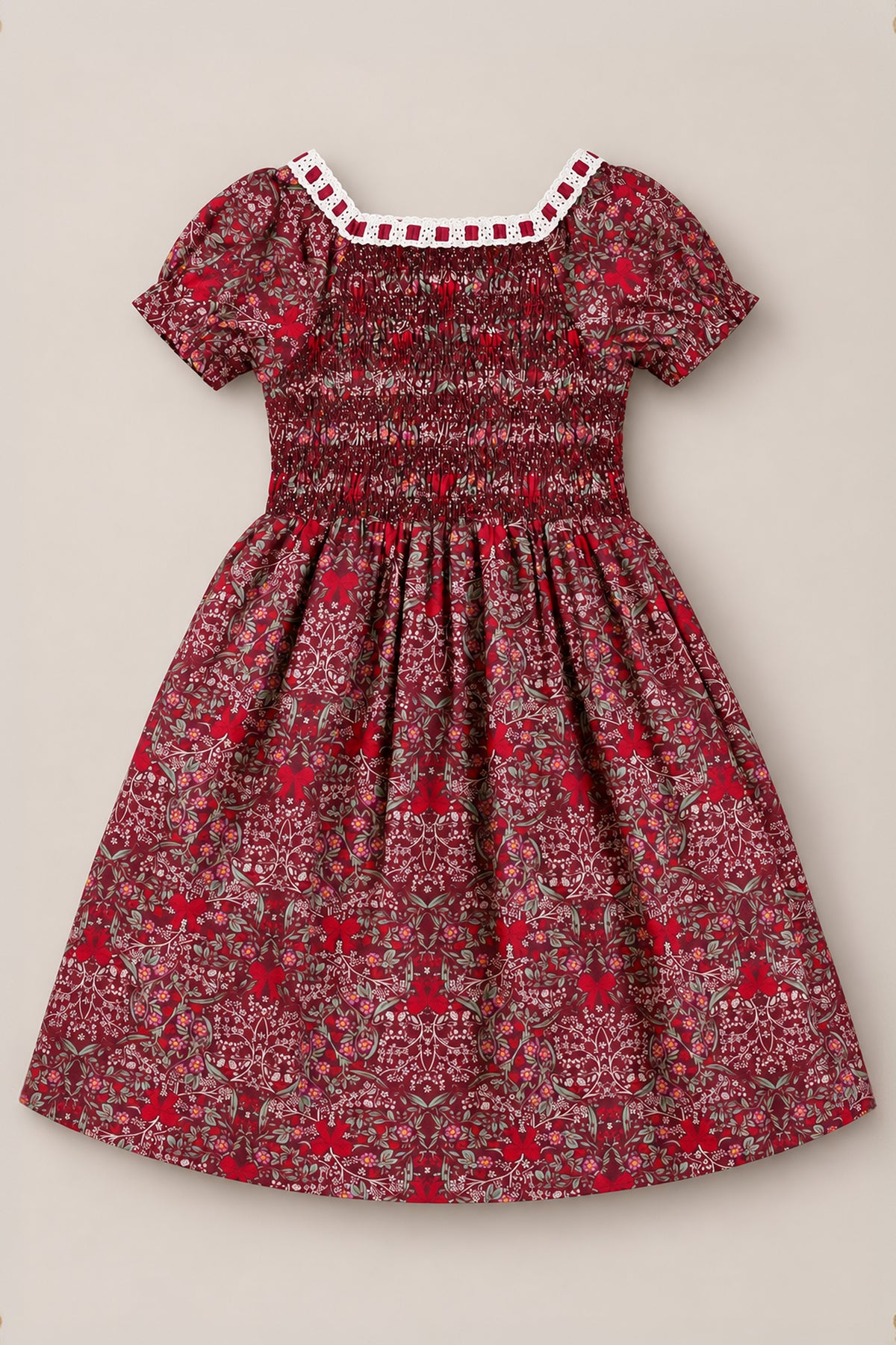 The Ivy City Co Mini Sunday Best Dress in Garnet Ribboned Garden is a burgundy girls’ holiday dress with short sleeves, featuring a white lace-trimmed neckline, a smocked bodice, and an all-over pattern of small red, white, and green florals and foliage. The gathered skirt falls to knee length for a charming finish.
