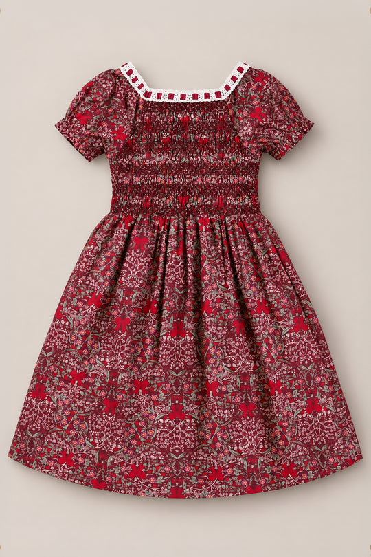 The Ivy City Co Mini Sunday Best Dress in Garnet Ribboned Garden is a burgundy girls’ holiday dress with short sleeves, featuring a white lace-trimmed neckline, a smocked bodice, and an all-over pattern of small red, white, and green florals and foliage. The gathered skirt falls to knee length for a charming finish.