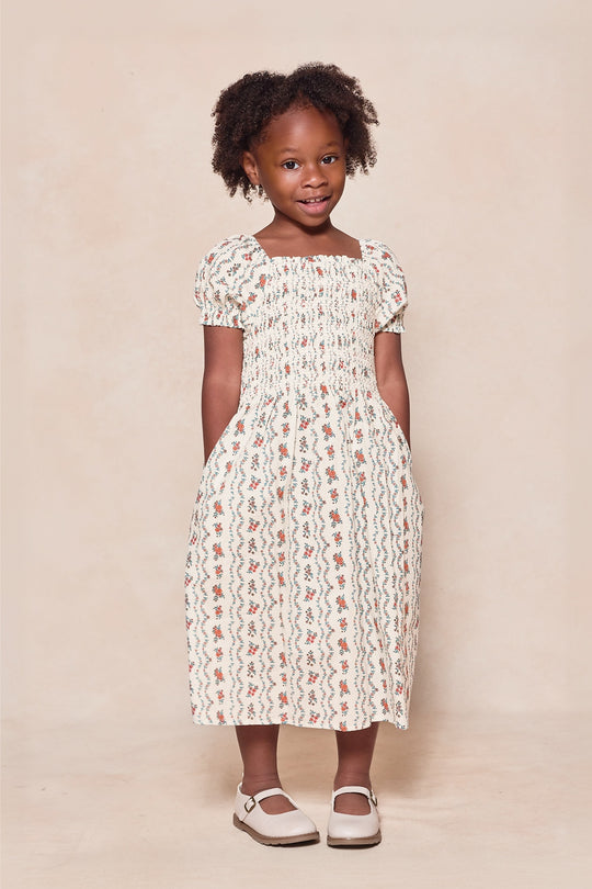 A young girl smiles while wearing the Ivy City Co Mini Sunday Best Dress in Linear Poppy Woodblock, a cream-colored, smocked cotton dress with a floral print. She has her hands in her pockets and stands against a light beige background.
