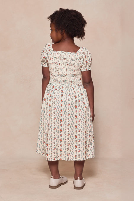 A young girl with curly hair faces away, wearing the Ivy City Co Mini Sunday Best Dress in Linear Poppy Woodblock—a white, calf-length summer dress with puff sleeves and a red and blue pattern—plus white shoes against a neutral background.