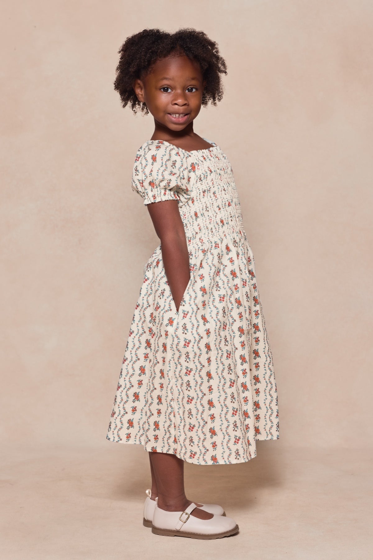 A young girl with curly hair smiles while posing sideways in the Ivy City Co Mini Sunday Best Dress in Linear Poppy Woodblock, a cream-colored, patterned cotton dress with short sleeves and white shoes against a neutral beige background.
