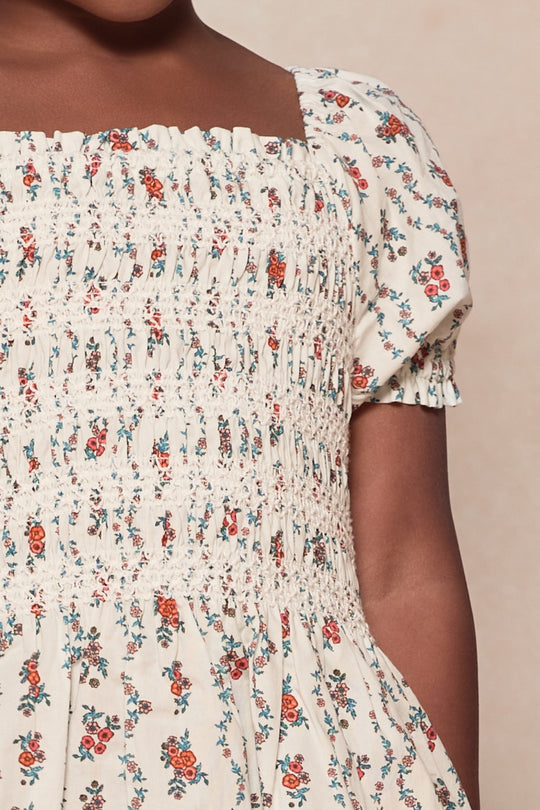 A close-up of the Mini Sunday Best Dress in Linear Poppy Woodblock by Ivy City Co—a cream cotton girls’ dress with red and blue floral pattern, smocked detail, and puff sleeves—perfect for sweet mom and me summer matching looks.