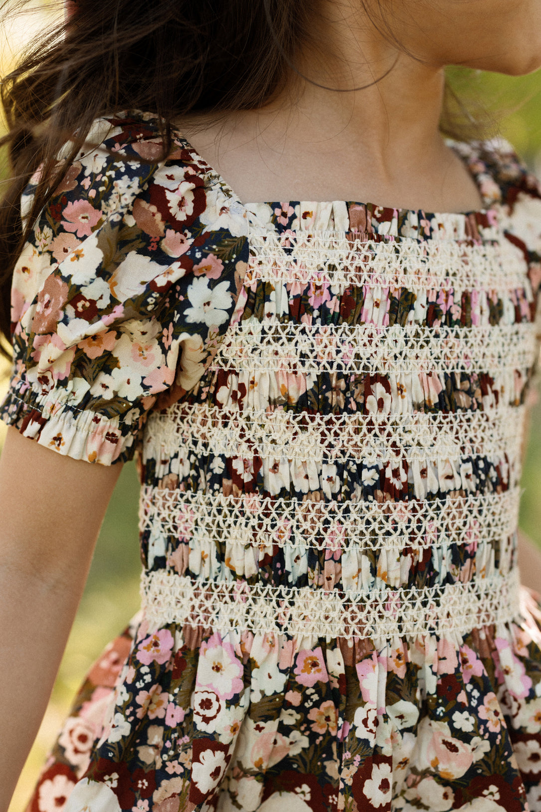 A child wearing the Ivy City Co Mini Sunday Best Dress in Midnight Garden, featuring puff sleeves, a floral pattern, and cream lace smocking on the bodice, stands outdoors with only the upper body and part of the face visible.