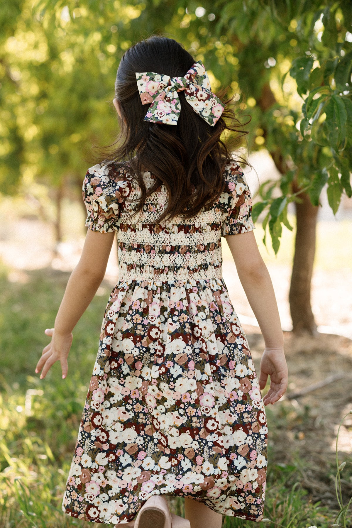A young girl with long hair, wearing the Ivy City Co Mini Sunday Best Dress in Midnight Garden—featuring a smocked bodice, puff sleeves, and a matching floral bow—walks outdoors among green trees on a sunny day.