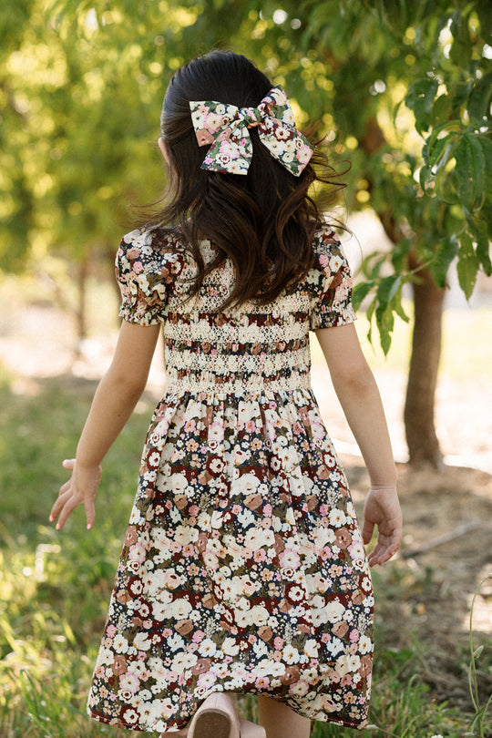 A young girl with long hair, wearing the Ivy City Co Mini Sunday Best Dress in Midnight Garden—featuring a smocked bodice, puff sleeves, and a matching floral bow—walks outdoors among green trees on a sunny day.