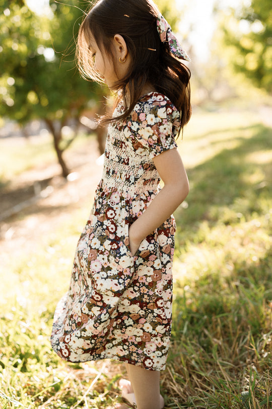 A young girl wearing the Ivy City Co Mini Sunday Best Dress in Midnight Garden, featuring puff sleeves, stands outdoors on the grass with sunlight illuminating her as she looks down, hands tucked into her pockets. Trees and greenery create a lush backdrop behind her.
