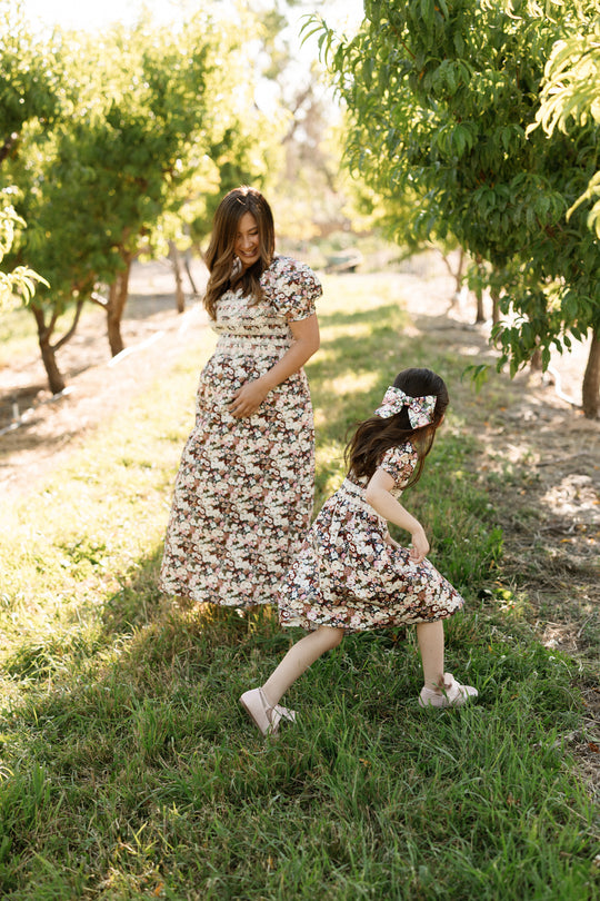 A woman and a young girl, both in Ivy City Co’s Mini Sunday Best Dress in Midnight Garden—featuring charming puff sleeves and a smocked bodice—are walking and playing together in a sunny orchard surrounded by lush green trees.