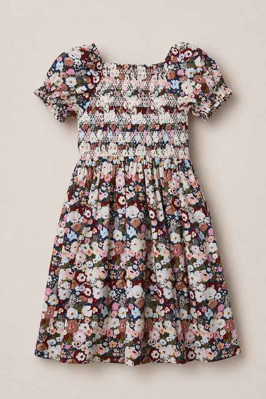 The Mini Sunday Best Dress in Midnight Garden by Ivy City Co is a short-sleeved, knee-length dress with a smocked bodice and puff sleeves, showcasing a colorful floral pattern in pink, purple, white, and green on a dark background.