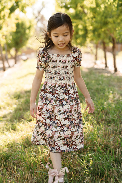 A young girl wearing the Ivy City Co Mini Sunday Best Dress in Midnight Garden, featuring a smocked bodice and puff sleeves, walks on grass in a sunlit outdoor setting with green trees in the background.