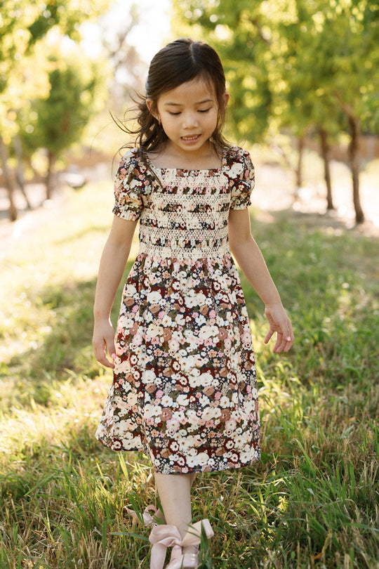 A young girl wearing the Ivy City Co Mini Sunday Best Dress in Midnight Garden, featuring a smocked bodice and puff sleeves, walks on grass in a sunlit outdoor setting with green trees in the background.