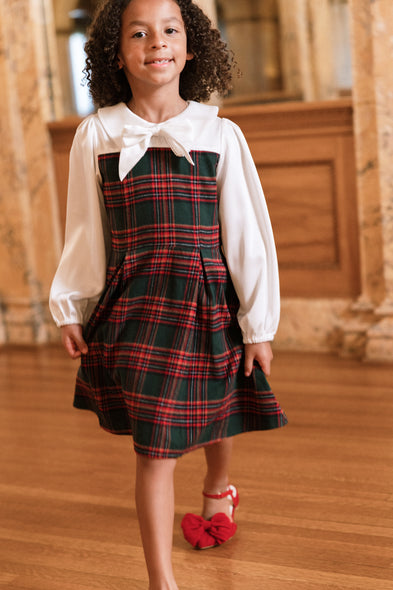 A young girl with curly hair, smiling at the camera as she walks indoors on a wooden floor, wears the Ivy City Co Mini Tawny Dress in Evergreen Plaid featuring a white collar and bow.