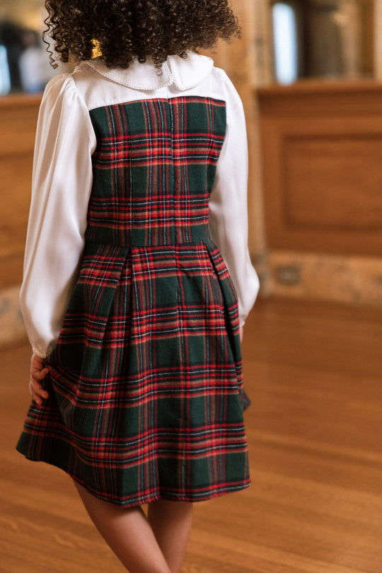 A child with curly hair stands indoors, facing away from the camera, wearing the Ivy City Co Mini Tawny Dress in Evergreen Plaid over a long-sleeve white blouse. The background features wooden paneling and a polished floor.