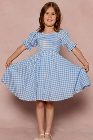 A smiling young girl poses against a pink backdrop, showing off the flared shape of her Ivy City Co Mini Teagan Dress in Blue Gingham—her loose brown hair and white shoes complete the charming girls’ spring dress look.