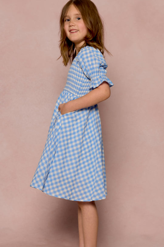 A young girl smiles sideways in the Ivy City Co Mini Teagan Dress in Blue Gingham, featuring short puffed sleeves and pockets, set against a soft pink background.