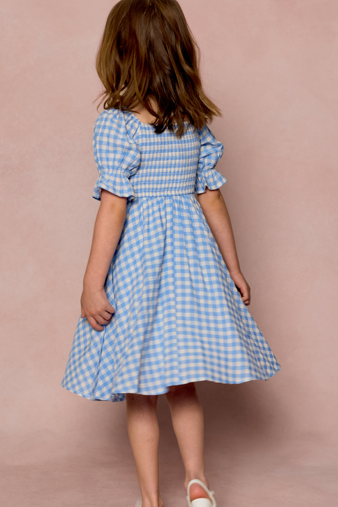 A young girl with brown hair, seen from behind, wears the Ivy City Co Mini Teagan Dress in Blue Gingham paired with white shoes, set against a soft pink background.