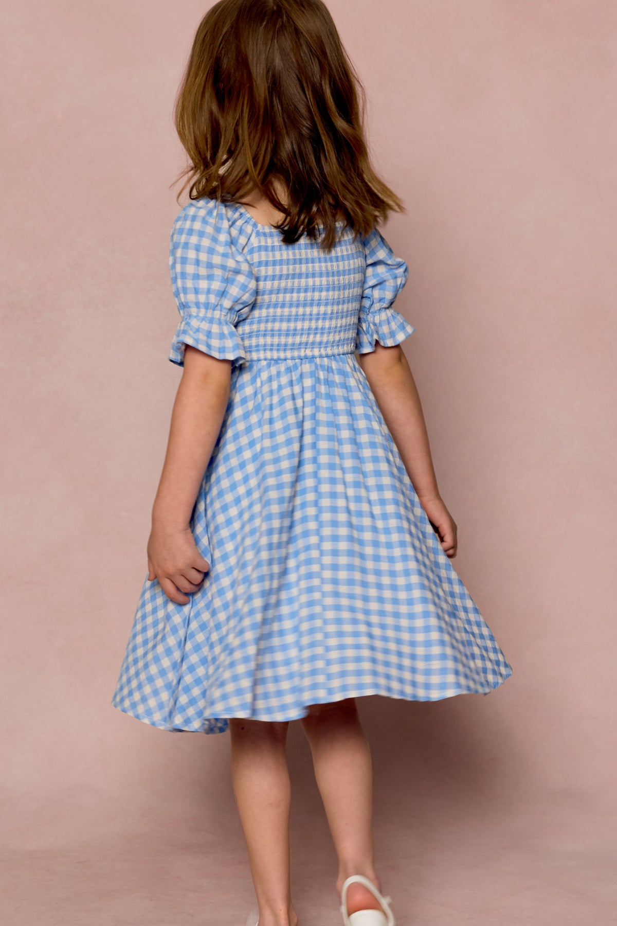 A young girl with brown hair, seen from behind, wears the Ivy City Co Mini Teagan Dress in Blue Gingham paired with white shoes, set against a soft pink background.