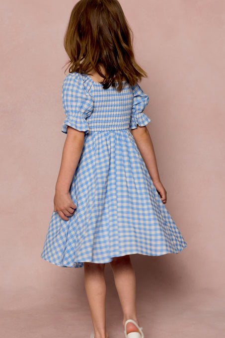A young girl with brown hair, seen from behind, wears the Ivy City Co Mini Teagan Dress in Blue Gingham paired with white shoes, set against a soft pink background.
