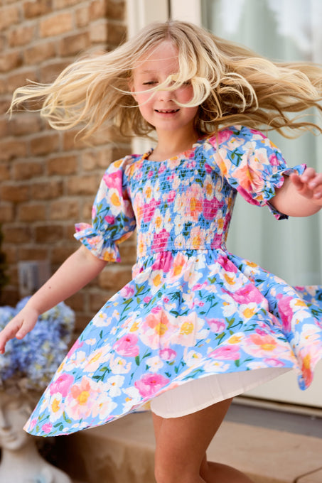A joyful young girl with blonde hair twirls outside in the Mini Teagan Dress in Delia Blue by Ivy City Co, a colorful floral cotton dress with puffed sleeves, her skirt and hair flowing as she dances near a brick wall.